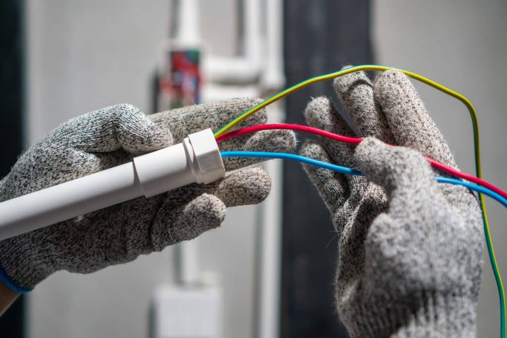 Electrician wearing protective gloves connects red, blue, and yellow-green electrical wires to a white conduit during an electrical installation.