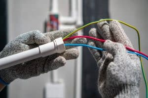 Electrician wearing protective gloves connects red, blue, and yellow-green electrical wires to a white conduit during an electrical installation.