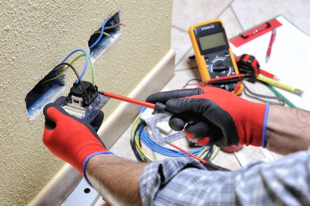 Electrician in protective gloves working on exposed electrical wiring with pliers.