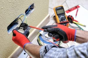 Electrician in protective gloves working on exposed electrical wiring with pliers.