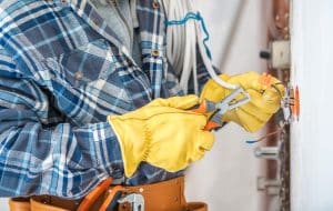 Electrician wearing yellow gloves uses pliers to repair exposed electrical wires inside a wall outlet.
